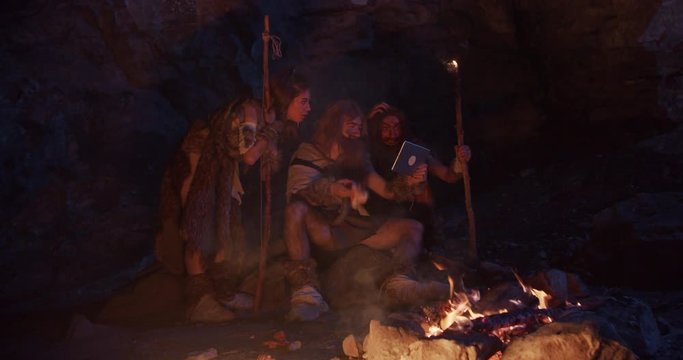 Tribe neanderthals wearing animal skin use tablet computer hold bone with meat having fun sitting by the fire in a cave stone ancient history evolution nature homo sapiens primitive campfire