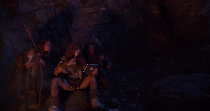 Close up cavemen hold bone with meat neanderthals wearing animal skin use tablet computer waving his hand having fun sitting by the fire in a cave stone ancient history evolution nature homo sapiens
