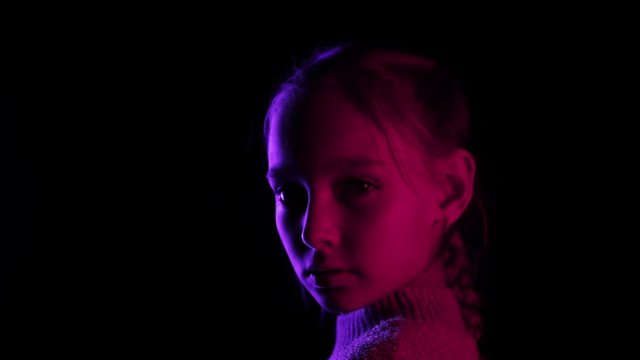 Teenager girl turning around on black background in neon lighting. Portrait young girl looking to camera in dark studio in blue and red lighting
