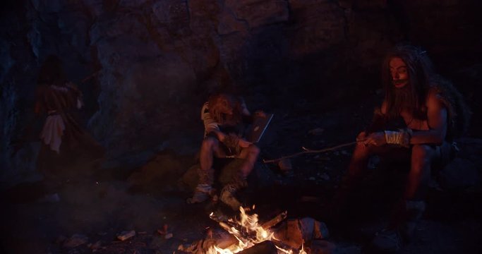 Neanderthals wearing animal skin discovering tablet computer sitting by the fire in a cave stone fun male ancient history portrait evolution nature homo sapiens primitive campfire slow motion