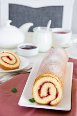 Homemade sponge cake with raspberry jam on a white plate on a white wooden dining table with a cup of fragrant berry tea.