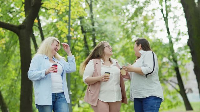 Three quarter length medium shot of group of three middle aged plus size women walking in green park with takeaway coffee cups and having joyful talk