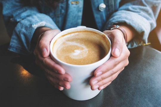Closeup image of a woman holding a cup of hot latte coffee on the table