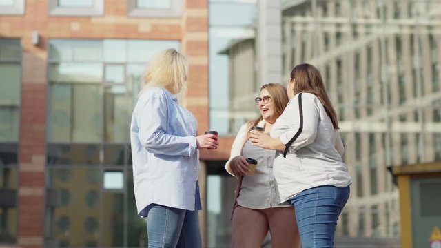 Three quarter length lockdown shot of corpulent woman joining two female friends talking over coffee in street and greeting them with embrace. Three oversize female friends chatting outdoors
