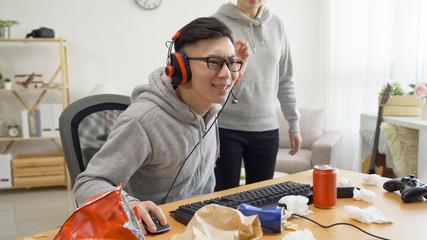 asian young man in headset playing online video game on computer intense concentrated. unrecognized angry mad annoyed girlfriend talking to him standing beside in dirty apartment. male ignored girl