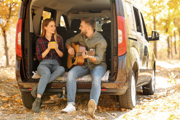 Young couple with guitar sitting in open car trunk outdoors