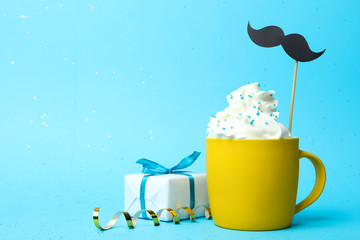 Cup of creamy drink with mustache and gift box on blue background, space for text. Greeting card