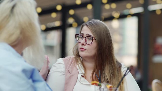 Sad plus size woman in eyeglasses telling female friend about her problems sitting at cafe table. Woman comforting girlfriend stroking her shoulder over cocktails