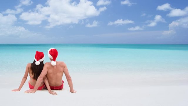 CINEMAGRAPH - seamless loop: Christmas couple sitting down relaxing on white sand beach sunbathing in tropical travel destination in winter holidays. Young adults wearing santa hat looking at view.