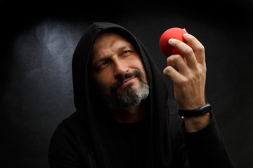 Portrait of a pensive bald man with a beard in a black hood with a red Christmas ball on a dirty gray background. Concept of young santa.
