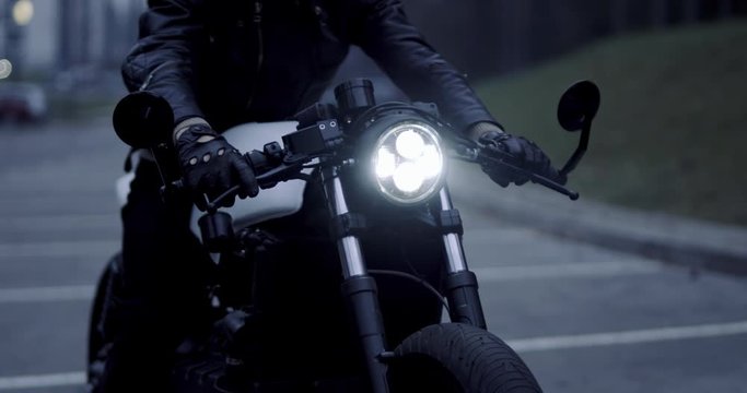 CU on headlight, Caucasian male biker wearing leather jacket sits on his custom built vintage cafe racer motorcycle at dawn. 4K UHD 50 FPS SLOW MOTION