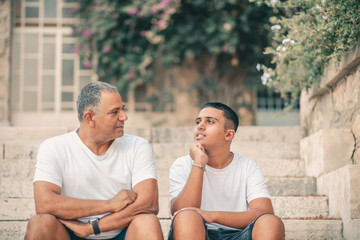Teenager son and senior father sitting on stairs outdoors at home, talking.
