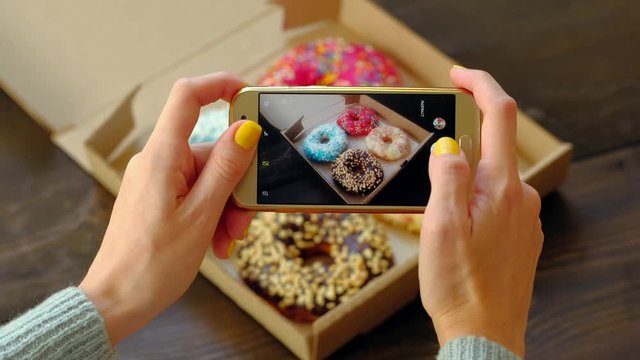 Woman's hands take pictures with smartphone of delicious fresh donuts in box on wooden table. 4k. Concept of food photo blogging.