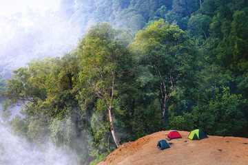 Adventure Camping Ground At The Edge of Top Mountain Cliff with Red, Blue and Green Tent During Sunrise with Fog Still Lingering on Rainforest or Jungle Wilderness Below.