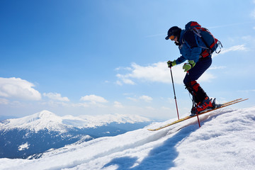 Fototapeta na wymiar Sportsman skier in skiing equipment with backpack jumping in air down steep snowy mountain slope on copy space background of blue sky and highland landscape. Winter sports, courage and speed concept.