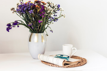 bouquet of flowers, open book and glasses, lemon tea in a white ceramic cup on a wicker stand on the table, cozy home interior.