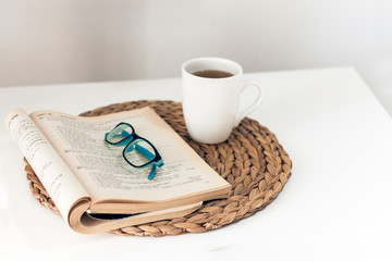 open book and glasses, lemon tea in a white ceramic cup on a wicker stand on the table, cozy home interior.