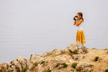 A girl with binoculars in her hands stands on Lake Baikal on the rocks and looks over the binoculars. Big stones. T-shirt and colored yellow skirt. Selective focus on the girl.
