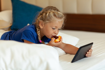 Little pretty girl holding tablet in her hands eats fruit and lies on bed (sofa) at home. Mobile game, entertainment and communication via Internet.
