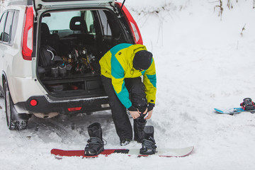 man changing regular boots to snowboard at parking place near car