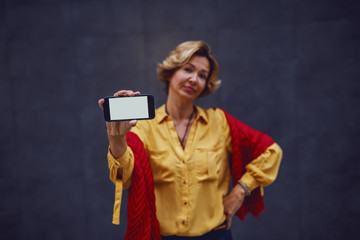 Attractive fashionable caucasian apathetic senior woman holding smart phone while standing with hand on hip. Selective focus on hand with smart phone.