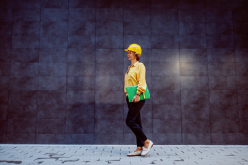 Full length of successful caucasian blonde senior female architect with helmet on head and folder with documents under armpit passing by gray wall and going to the construction site. © Dusan Petkovic