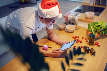 Handsome caucasian smiling chef in white uniform and with santa hat on head using tablet while...
