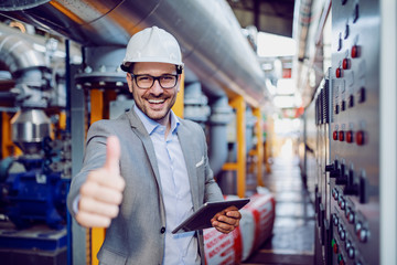 Successful caucasian supervisor in suit and with helmet holding tablet and showing thumbs up while standing next to dashboard in power plant. - obrazy, fototapety, plakaty