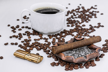 On a white table among coffee beans in the foreground lies a cigar on an ashtray, next to a lighter. In the background a defocused white cup of black aromatic coffee