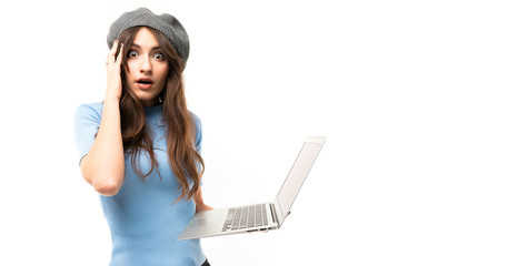 Young girl with delightful smile, long wavy chestnut hair, beautiful makeup, in blue jersey, black jeans, grey beret, standing with laptop and is shocked