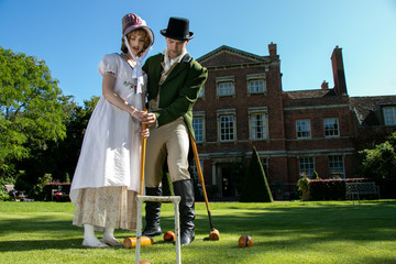Handsome man and beautiful woman dressed in vintage clothing on lawn in front of stately home,...