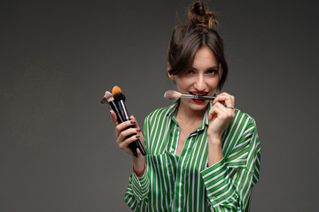 A young beautiful woman with long wavy chestnut hair stuffed in a sloppy bundle, strands on the sides of her face, bright makeup, in a white-green striped shirt, holds makeup brushes