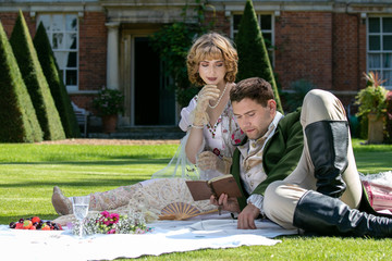 Young lovers dressed in vintage clothing sitting on picnic blanket. Gentleman reading to his lover from a book of poems