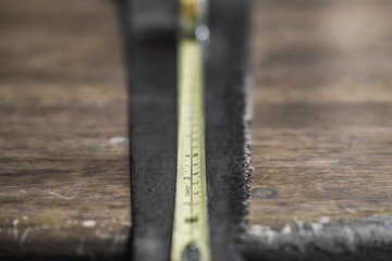 Close-up, shallow focus of an extended measuring tape showing the scales. Also are an old hammer and wood saw seen on an old, wooden workshop table.