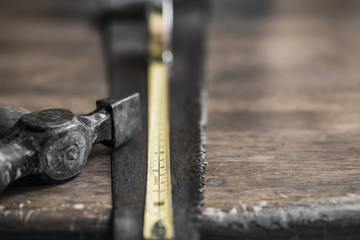 Close-up, shallow focus of an extended measuring tape showing the scales. Also are an old hammer and wood saw seen on an old, wooden workshop table.