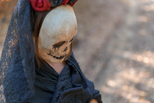 Close up photo of woman with Halloween mask in autumn woods. The girl is wearing black costume and mask with flowers typical for celebrating the  Mexican holiday Day of the dead.