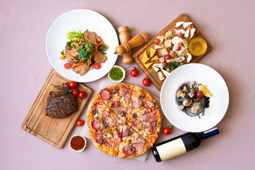 Dining table with food, top view. Pizza, salad, cheese board, steak, black seafood pasta, bottle of wine.