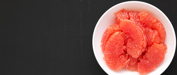 Red grapefruit slices in a white bowl on a black background, top view. Flat lay, overhead, from above. Space for text.