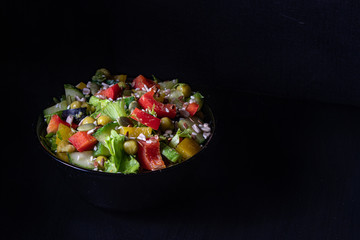 Vegetable salad in a dark bowl on a dark background. Green Leaf Lettuce, cucumber, bell pepper, green peas, black and green olives, sprinkled with sunflower seeds, pumpkin seeds, sesame seeds.