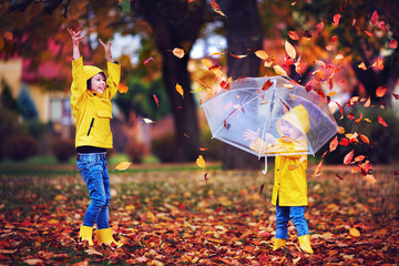 happy excited kids having fun, throwing leaves in autumn park, wearing rain boots and coats
