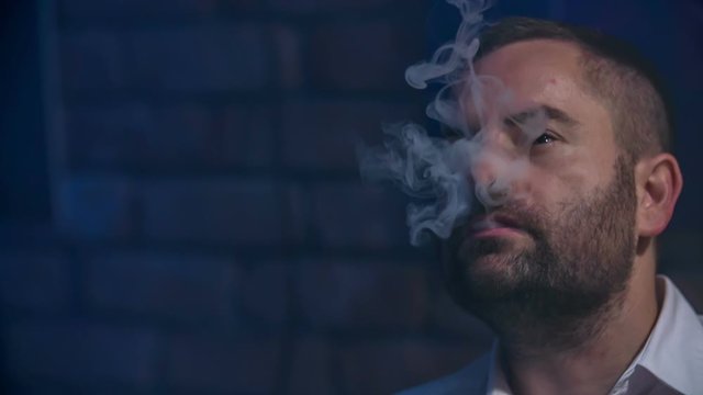 This man is really slowly smoking in a bar and he is letting the smoke out of his mouth.