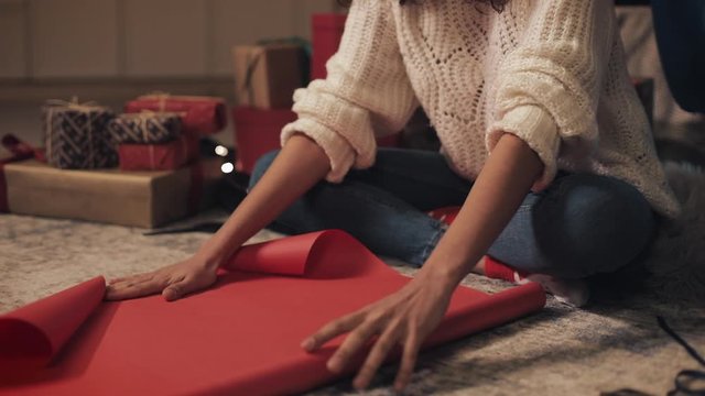 Close Up of Young Beautiful Girl Sitting under Decorated Cristmas Tree, Preparing Presents Deploying the Scroll of Paper, Putting Present Box on it. Holiday Celebration Concept.