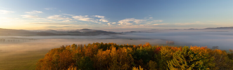 Fototapeta na wymiar Aerial panoramic view to autumn foliage trees and meadow with misty fog and hill at sunrise, Czech landscape