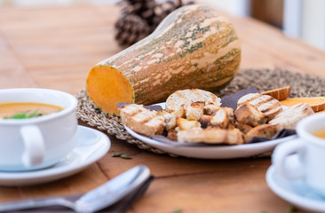 Wooden rustic table with white bowls of pumpkin cream soup. In the middle of the wooden table bread croutons and the raw vegetable