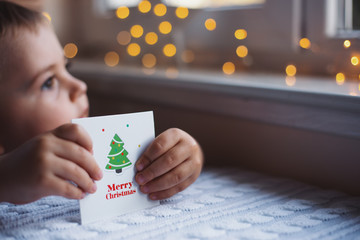 Child boy holding Christmas greeting card with warm garland bokeh on background near window in...