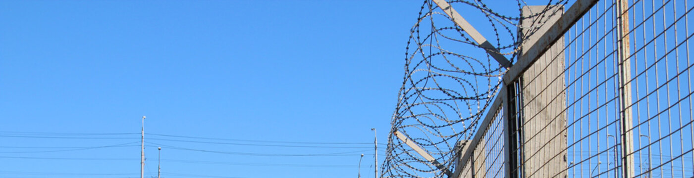 Barbed Wire On The Fence, Concept For The Protection Of Territory, Property, Military Installations, The US And Mexico Border Or Places Of Detention, Wide View Against A Blue Sky.