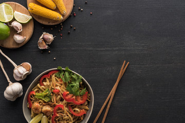 top view of tasty spicy thai noodles with shrimps near chopsticks and fresh ingredients on wooden grey surface with copy space