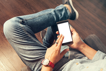 Man in bluejeans wearing a red watch and a ring is using a smart phone seated with crossed legs,...