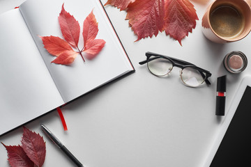 top view of notebook near glasses, coffee cup, cosmetics, pen and red leaves of wild grapes on white table