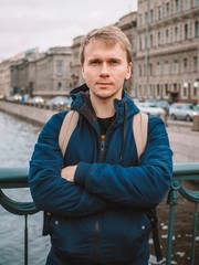 A blond man in a jacket and with a backpack stands on the bridge against the background of the city of St. Petersburg on an autumn cloudy day